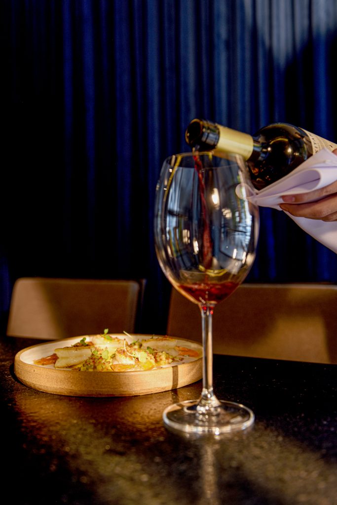 Taça de vinho sendo servida com vinho vermelho, com um prato de refeição posto na mesma mesa.