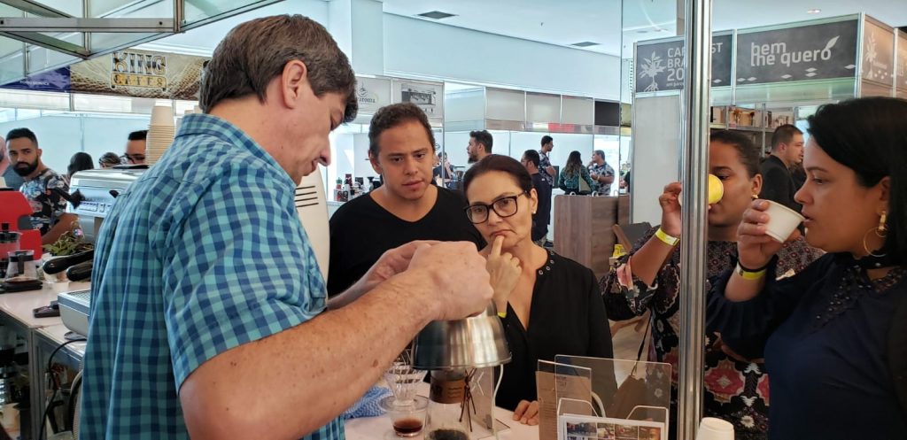 Na imagem, barista demonstra técnicas de café em estande para um grupo de 4 participantes do evento. 