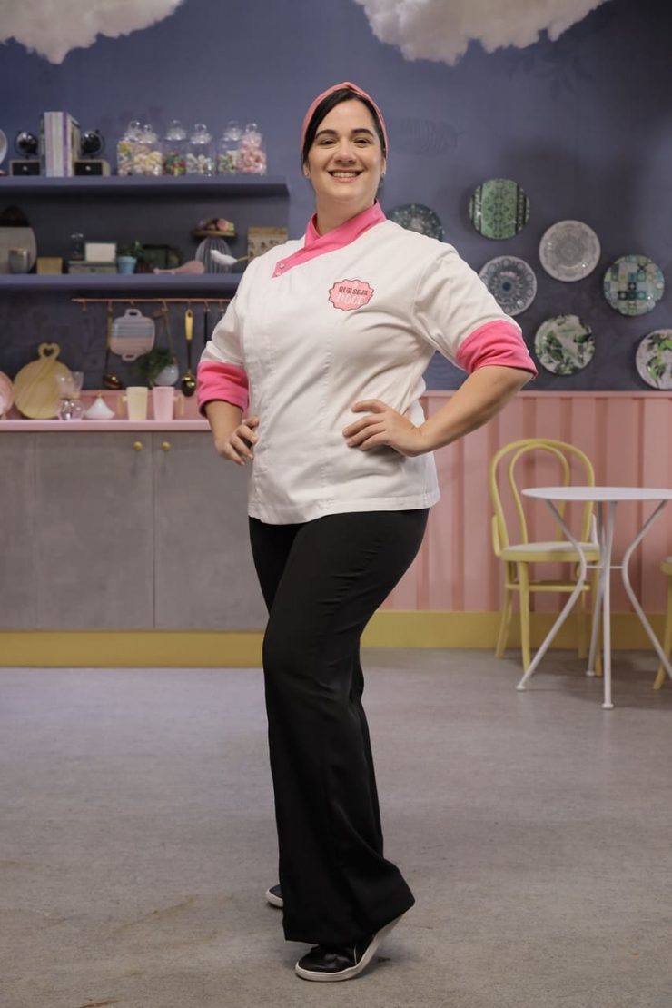 Mari posa com o traje do programa, nas cores Branco com Rosa. Ao fundo, o cenário de gravação, com predominância de tons roxos e pasteis.